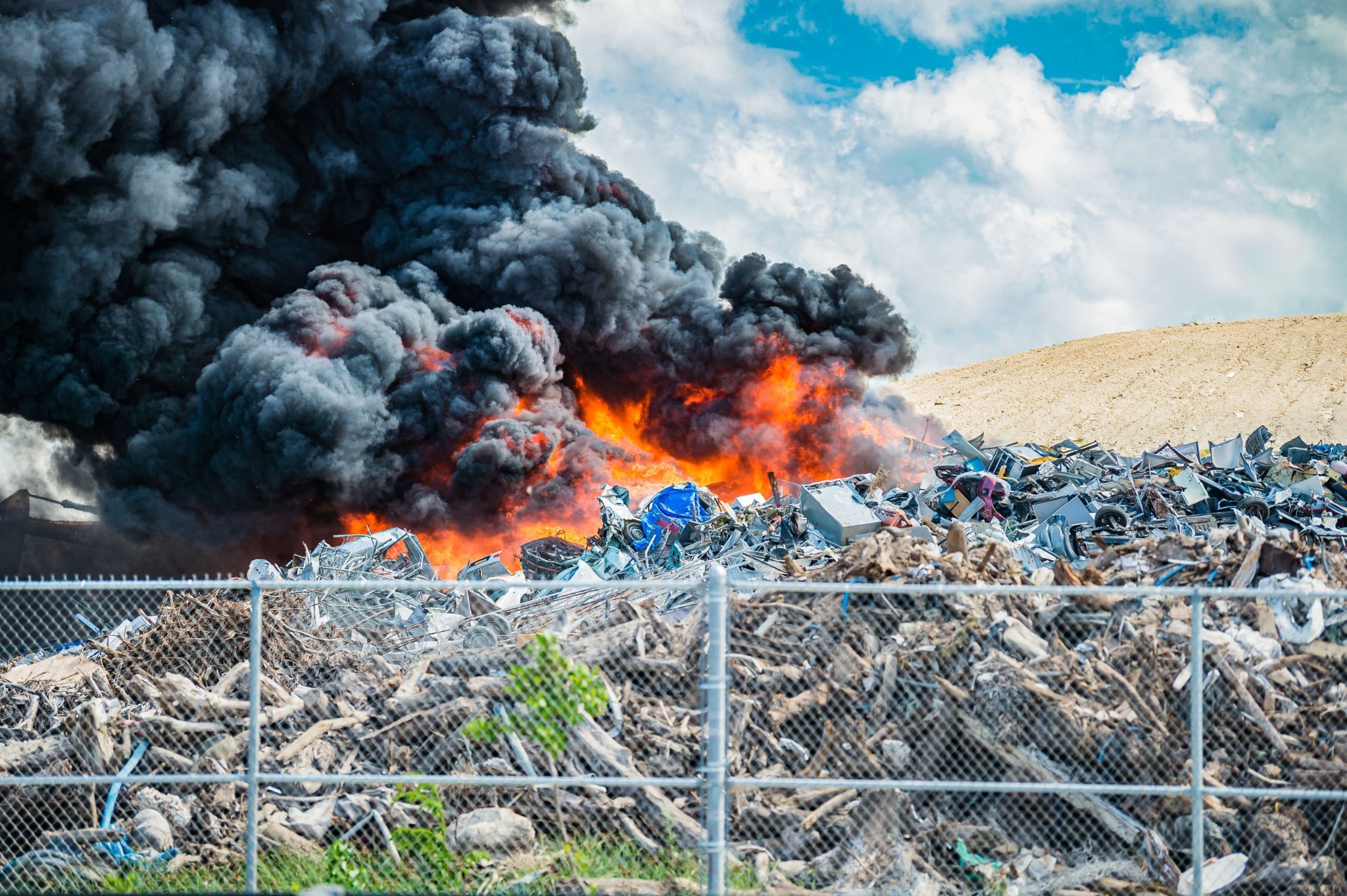 Flames and a dense dark plume rising behind a yard of discarded materials.
