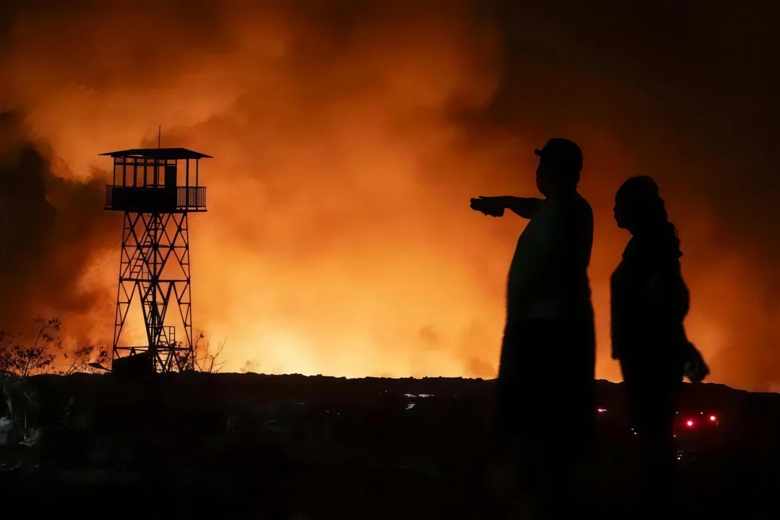 Two residents and a watchtower silhouetted against orange smoke from a night-time fire at Jatibarang landfill